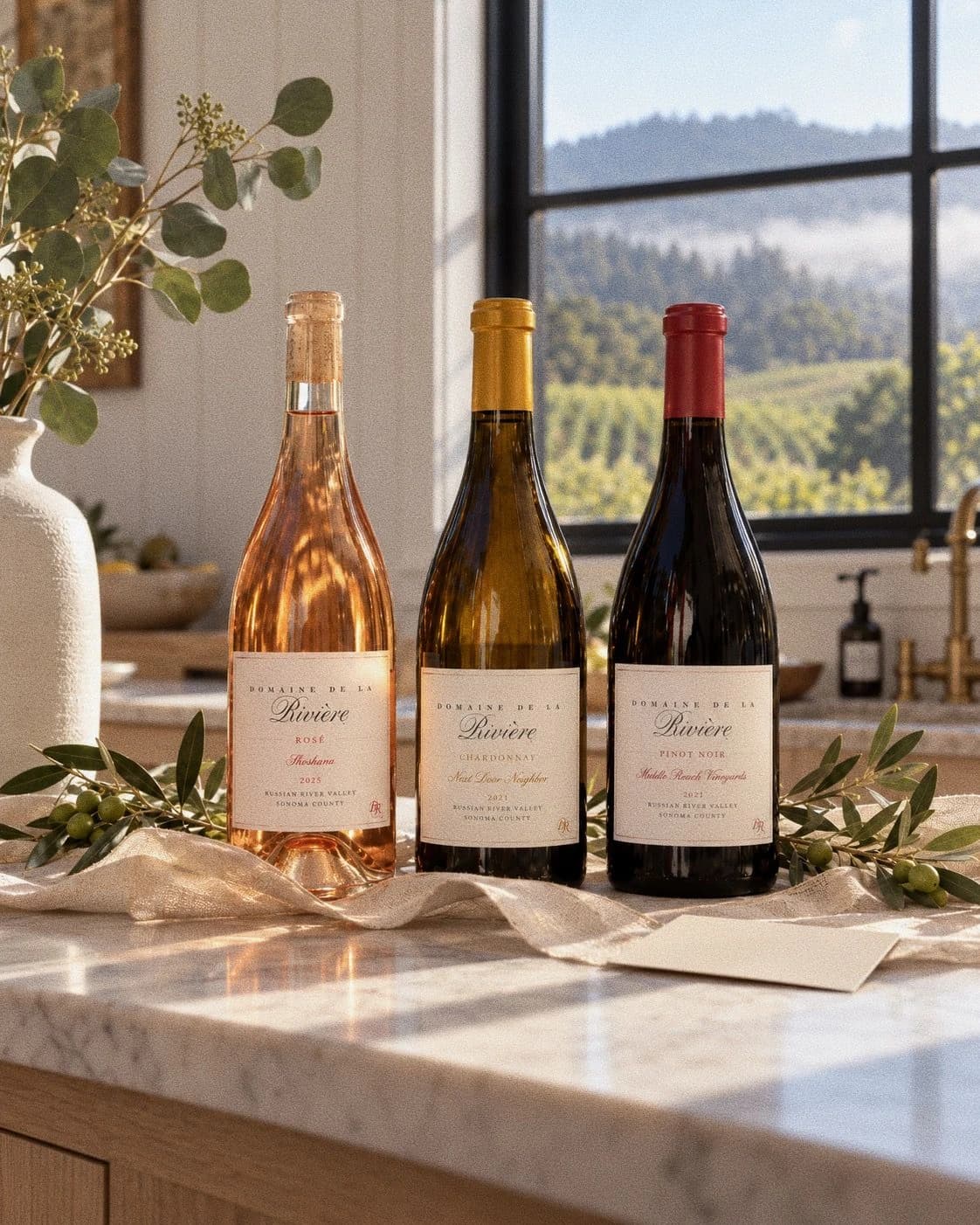 2025 Shoshana Rosé, 2021 Next Door Neighbor Chardonnay, and 2021 Middle Reach Vineyards Pinot Noir on a marble kitchen counter by a vineyard window
